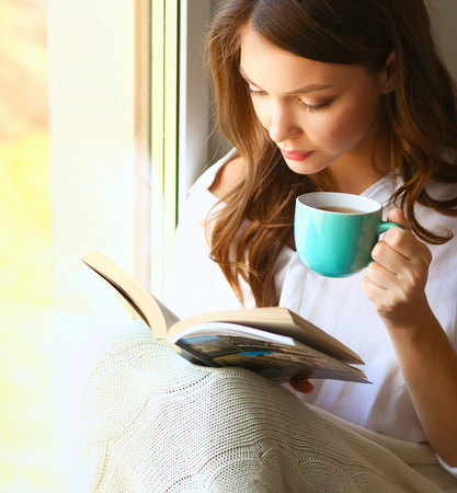 Young woman at home sitting near window relaxing in her living room reading book and drinking coffee or tea .の写真素材