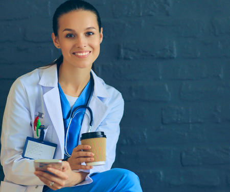Female doctor sitting with mobile phone and drinking coffeeの写真素材