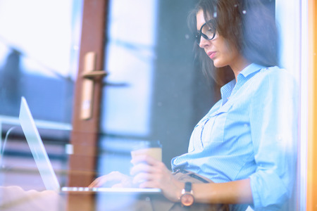 Young woman sitting at office table with laptop,view through windowの写真素材