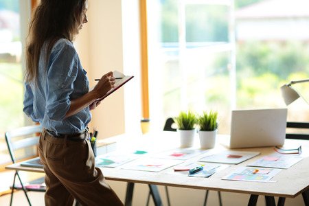 Young woman standing near desk with instruments, plan and laptopの写真素材