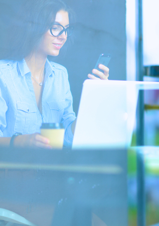 Beautiful young business woman sitting at office desk and holding cell phone,view through windowの写真素材