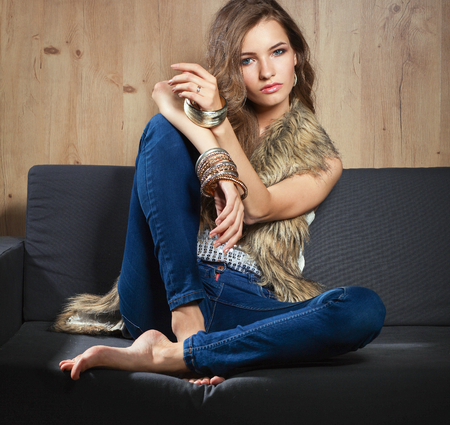 Portrait of a elegant woman sitting on a black sofa wearing a blue jeans and fur vestの写真素材