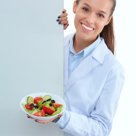 Portrait of a beautiful woman doctor holding a plate with fresh vegetables standing near blankの写真素材
