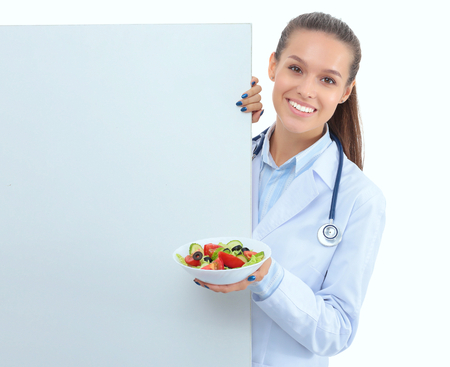 Portrait of a beautiful woman doctor holding a plate with fresh vegetables standing near blankの写真素材