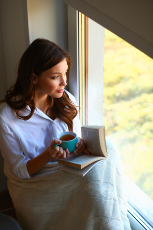 Young woman at home sitting near window relaxing in her living room reading book and drinking coffee or teaの写真素材