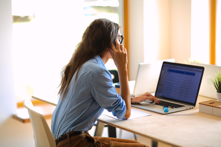 Beautiful young business woman sitting at office desk and talking on cell phoneの写真素材