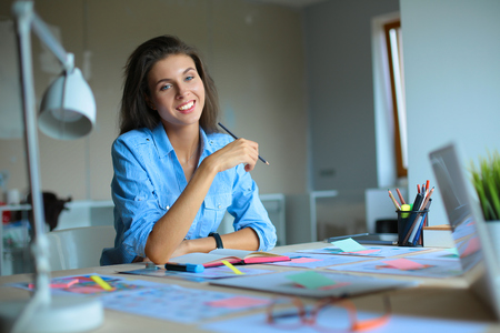 Young woman sitting at the desk with instruments, plan and laptopの写真素材