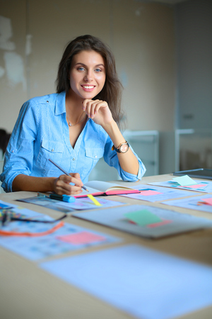 Young woman sitting at the desk with instruments, plan and laptopの写真素材