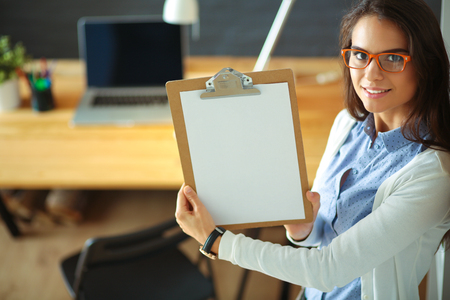 Young woman standing near desk with laptop holding folderの写真素材