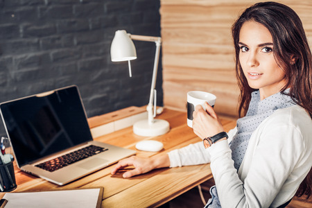 Portrait of relaxed young woman sitting at her desk holding cup of coffeeの写真素材
