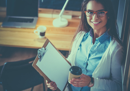 Young woman standing near desk with laptop holding folder and cup of coffeeの写真素材