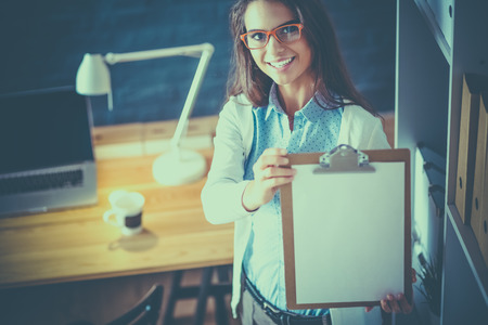 Young woman standing near desk with laptop holding folderの写真素材