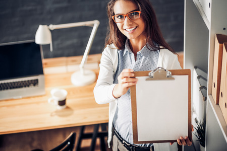 Young woman standing near desk with laptop holding folderの写真素材