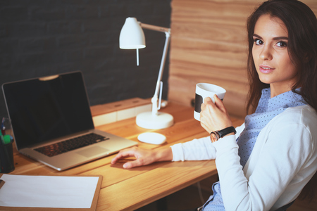 Portrait of relaxed young woman sitting at her desk holding cup of coffeeの写真素材