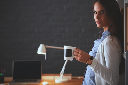 Young woman standing near desk with laptop holding cup of coffeeの写真素材