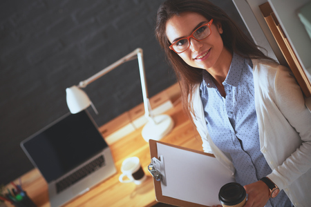 Young woman standing near desk with laptop holding folder and cup of coffeeの写真素材
