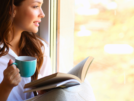 Young woman at home sitting near window relaxing in her living room reading book and drinking coffee or tea.の写真素材