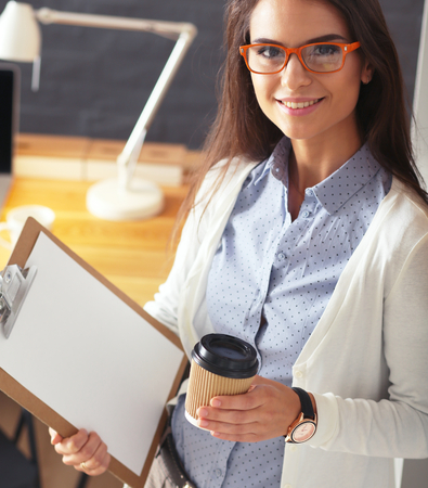 Young woman standing near desk with laptop holding folder and cup of coffeeの写真素材