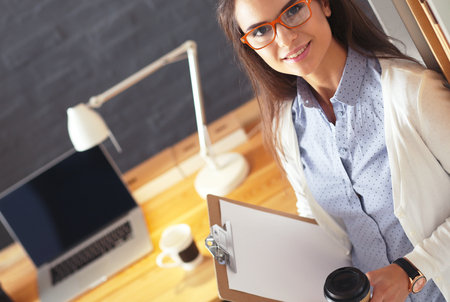 Young woman standing near desk with laptop holding folder and cup of coffeeの写真素材