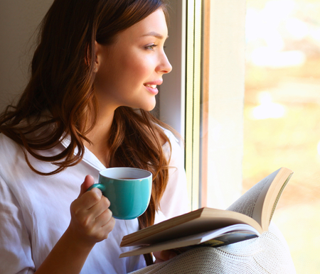 Young woman at home sitting near window relaxing in her living room reading book and drinking coffee or tea.の写真素材