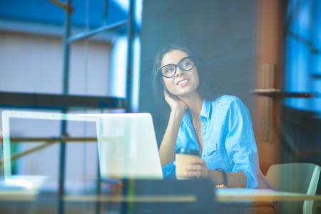 Young woman sitting at office table with laptop,view through windowの写真素材