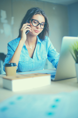 Beautiful young business woman sitting at office desk and talking on cell phone.の写真素材