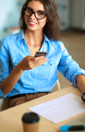 Beautiful young business woman sitting at office desk and holding cell phoneの写真素材