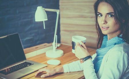 Portrait of relaxed young woman sitting at her desk holding cup of coffeeの写真素材
