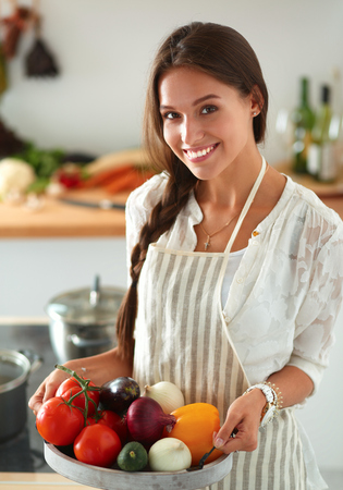 Smiling young woman holding vegetables standing in kitchenの写真素材