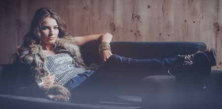 Portrait of elegant woman sitting on black sofa wearing a blue jeans and fur vestの写真素材