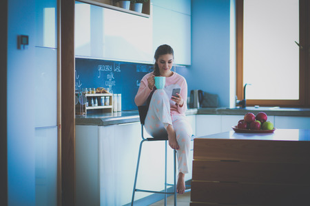 Woman using mobile phone sitting in modern kitchenの写真素材