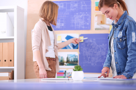 Two young woman standing near desk with instruments, plan and laptop.の写真素材