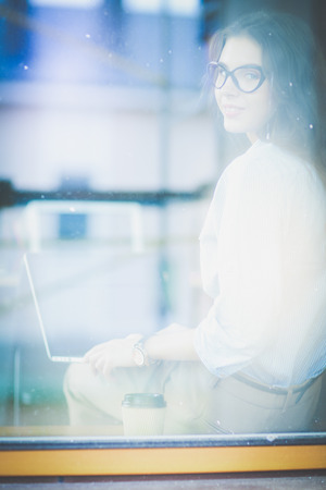 Young woman sitting at office table with laptop,view through window. Young womanの写真素材