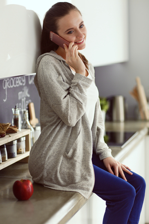 Woman using mobile phone sitting in modern kitchen.の写真素材
