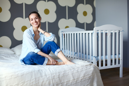 Young woman sitting on the bed near childrens cot. Young momの写真素材