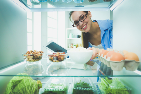 Portrait of female standing near open fridge full of healthy food, vegetables and fruitsの写真素材
