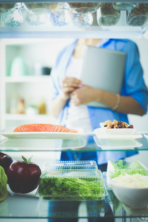 Portrait of female standing near open fridge full of healthy food, vegetables and fruitsの写真素材