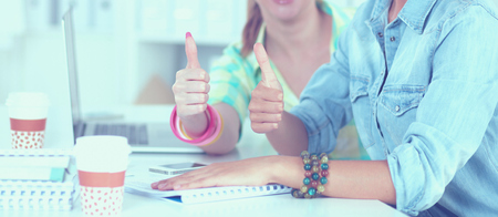 Two women working together at office, sitting on the desk and showing ok.の写真素材