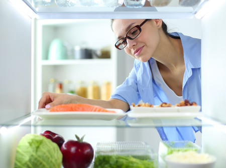 Portrait of female standing near open fridge full of healthy food, vegetables and fruits.の写真素材