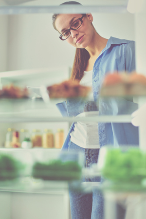 Portrait of female standing near open fridge full of healthy food, vegetables and fruits.の写真素材