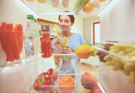Portrait of female standing near open fridge full of healthy foodの写真素材