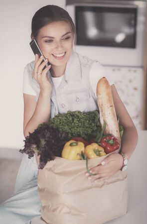Smiling woman with mobile phone holding shopping bag in kitchenの写真素材