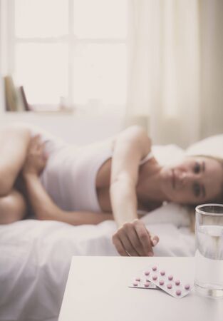 Young beautiful woman with pills lying on bed in bedroomの写真素材