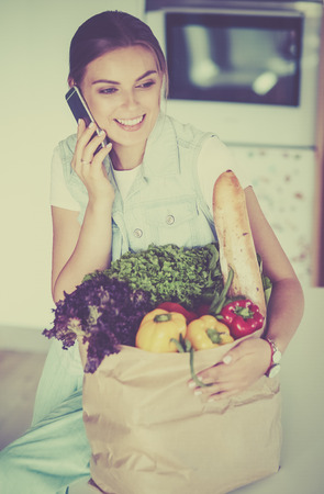 Smiling woman with mobile phone holding shopping bag in kitchenの写真素材