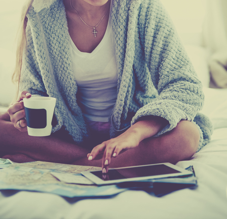 Relaxed young woman sitting on bed with a cup of coffee and digital tabletの写真素材