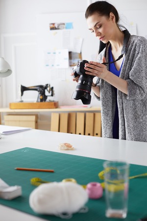 Young designer woman taking photos with digital camera while standing near the desk in her workshopの写真素材