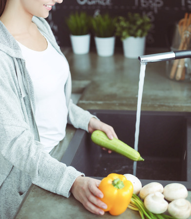 Beautiful young woman washing vegetables for salad while standing in the kitchen.の写真素材
