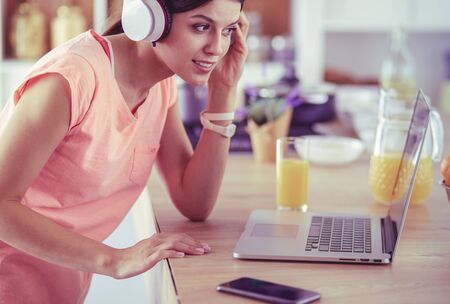 Portrait of a cheerful young woman listening to music with headphones and using laptop computer while standing at the kitchenの写真素材