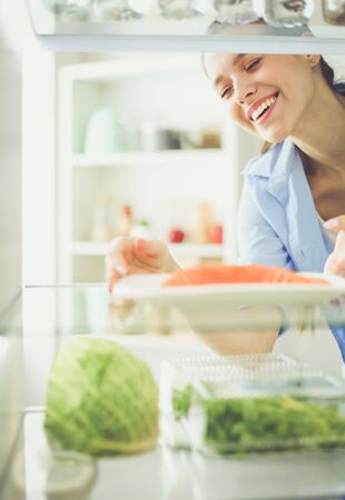 Portrait of female standing near open fridge full of healthy food, vegetables and fruits.の写真素材