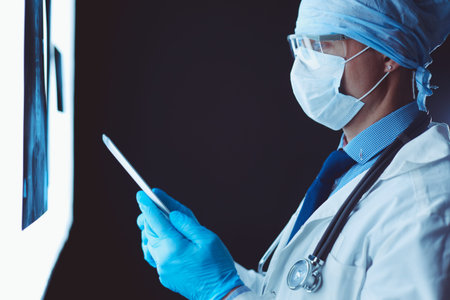 Doctor in hospital sitting at desk looking at x-rays on tablet against white background with x-rays.